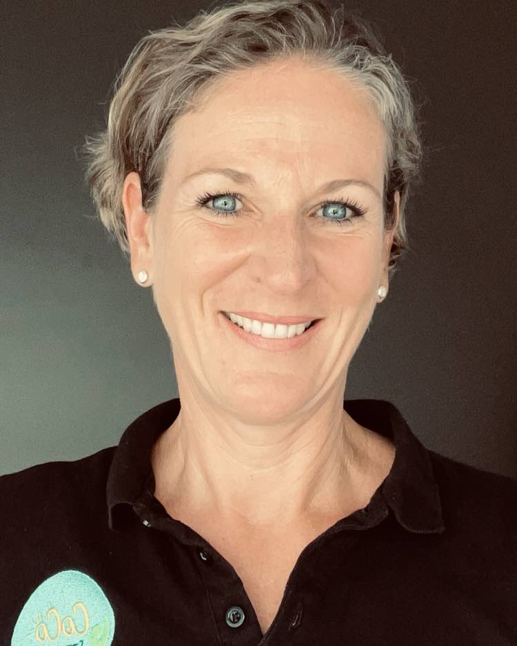 Corinna, a woman in her fifties with short grey hair, smiling directly at the camera in a black CoCo Fitness polo shirt with the embroidered badge on the chest.