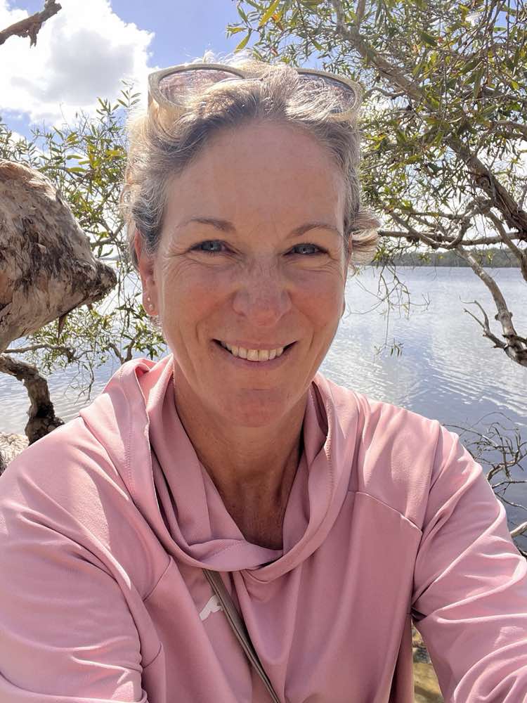 Corinna outdoors beside a Noosa lake in a pink hooded top, sunglasses pushed up into her hair, smiling at the camera under a paperbark tree.