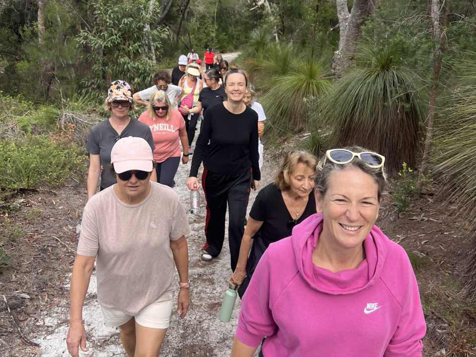 Corinna walking a sandy bush track in Noosa in a pink hoodie, smiling at the camera, with eight women of the Noosa Walk and Talk group following the trail behind her.